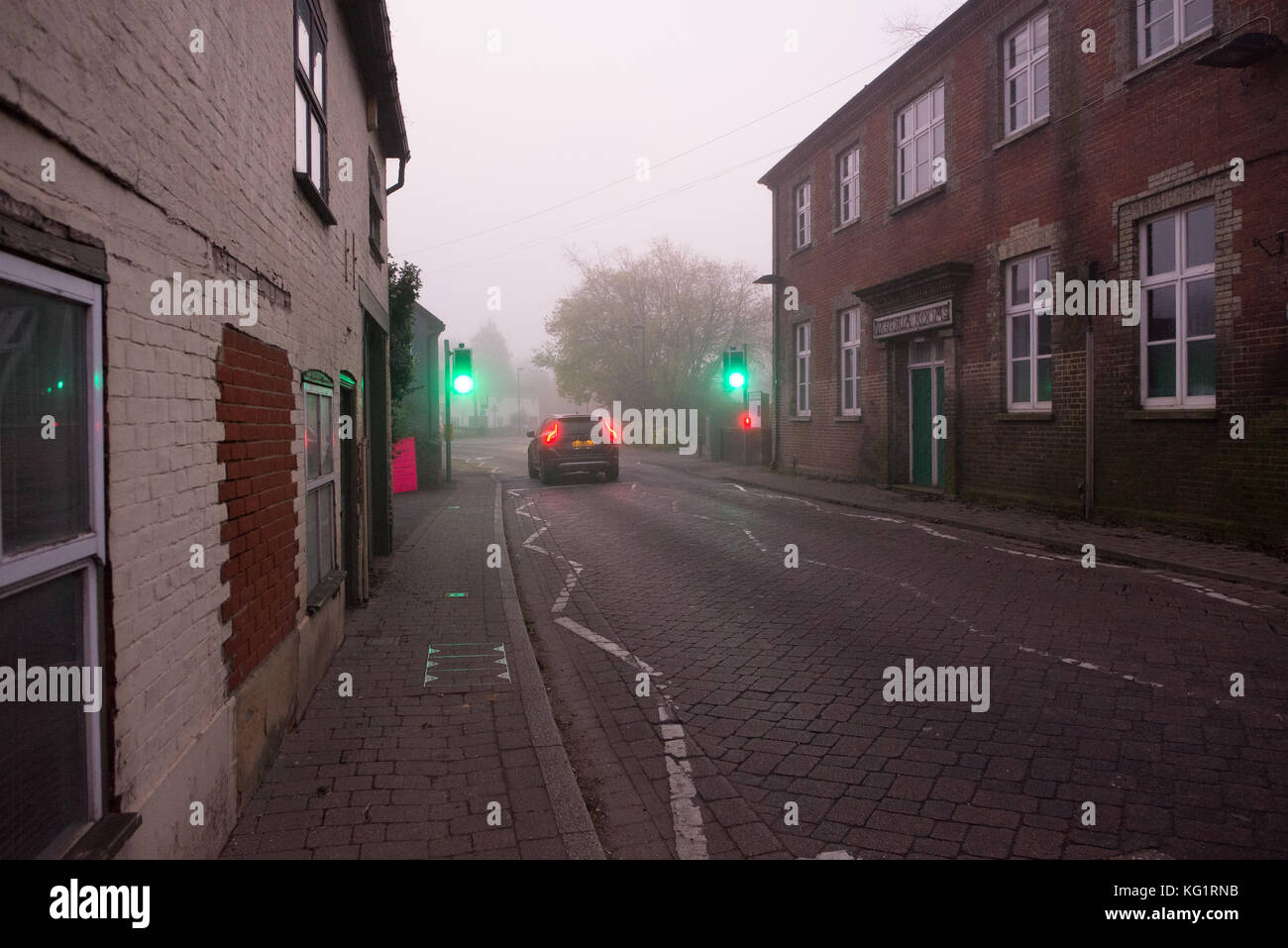 Foggy morning in the New Forest town, Bridge Street, Fordingbridge, Hampshire, UK, 3rd November 2017. Car passing through green traffic lights with fog lights on. Stock Photo