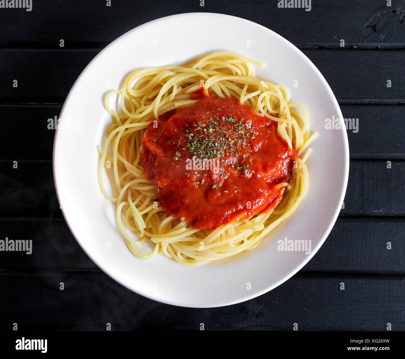 Cooked spaghetti pasta dish, hot with steam coming out, with red tomato sauce and oregano sprinkled on top, on a black wooden table background Stock Photo