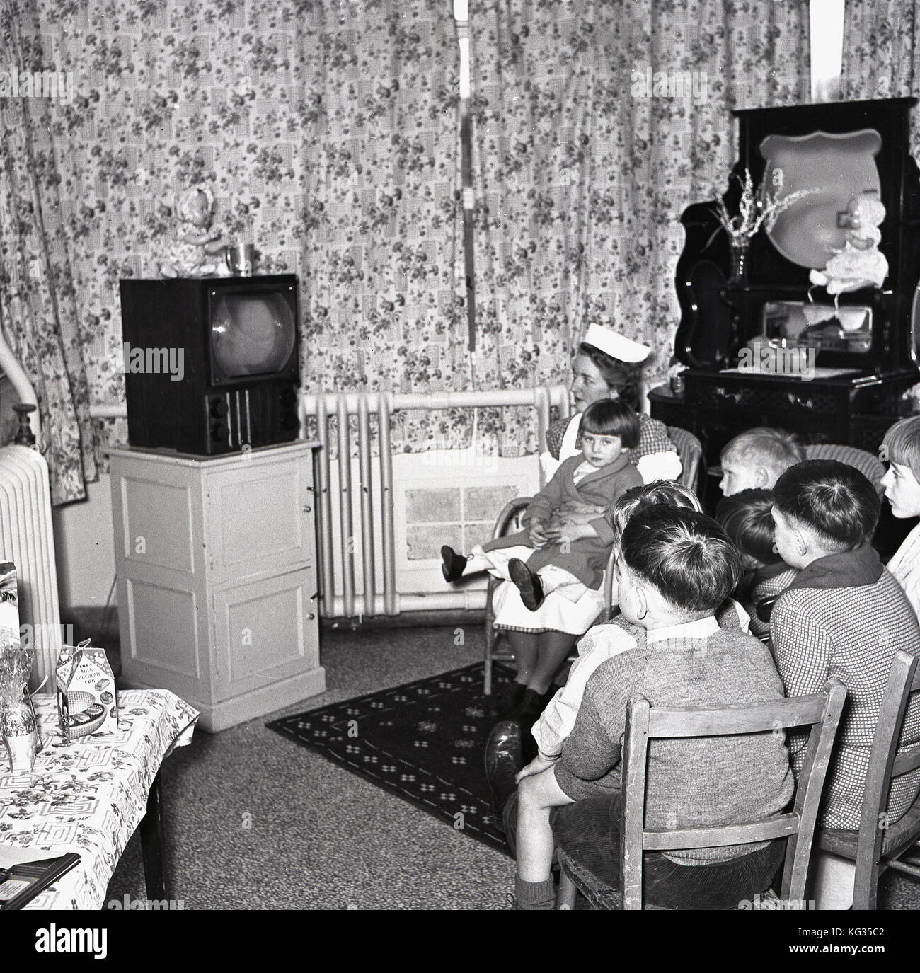 1950s, England, TV room in a children's hospital, group of small children sitting together watching a television, with one young girl held by a nurse. Stock Photo