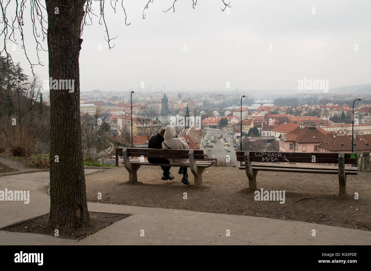 A view of Prague from a hill with benches in the foreground and a couple sitting on one on an overcast day. Stock Photo