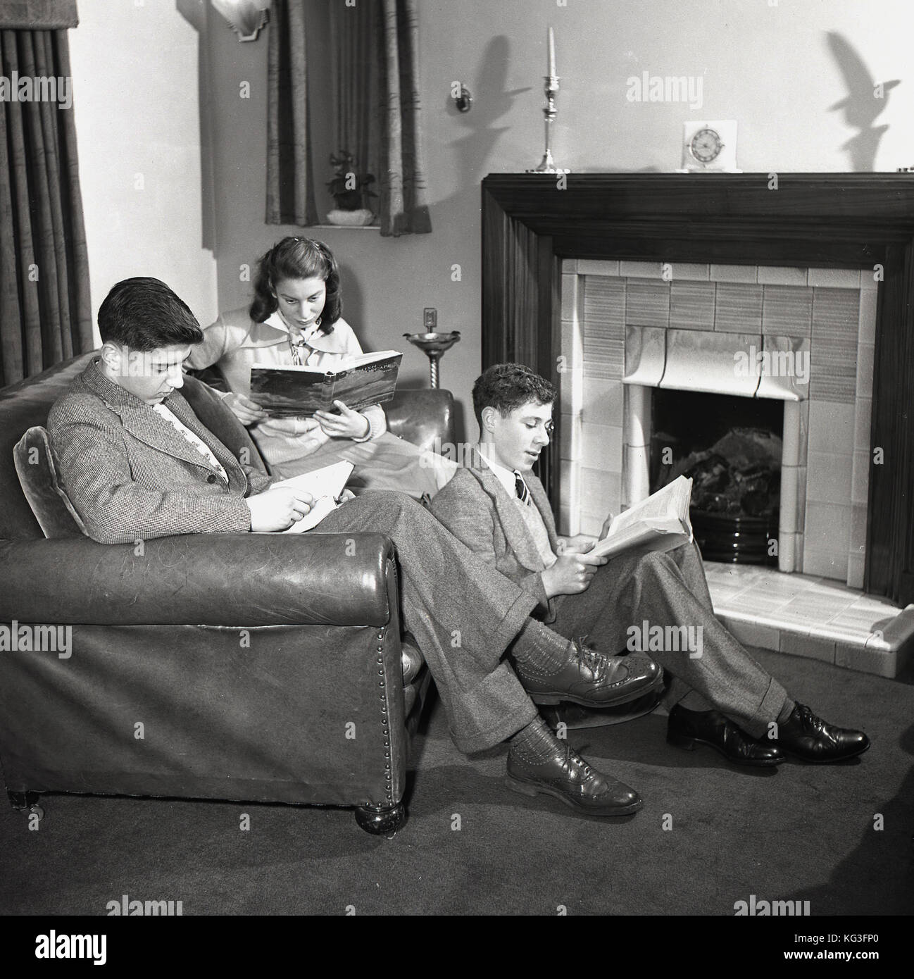 1950s, historical picture of three young adults in a front room, two sitting on a leather sofa, one sitting on the floor, reading books, England, UK. Stock Photo