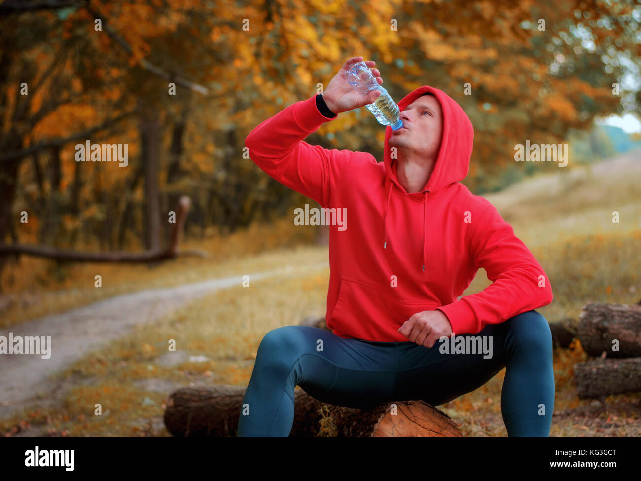 A young athletic runner in a red sports jacket with a hood and black sport leggins drink water from the bottle after jogging on a colorful autumn fore Stock Photo