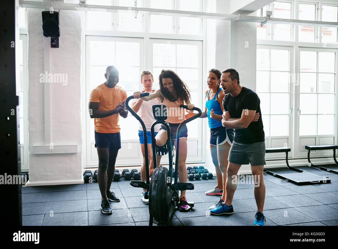 Group of smiling people cheering on their friend riding a stationary bike while working out together in a gym Stock Photo