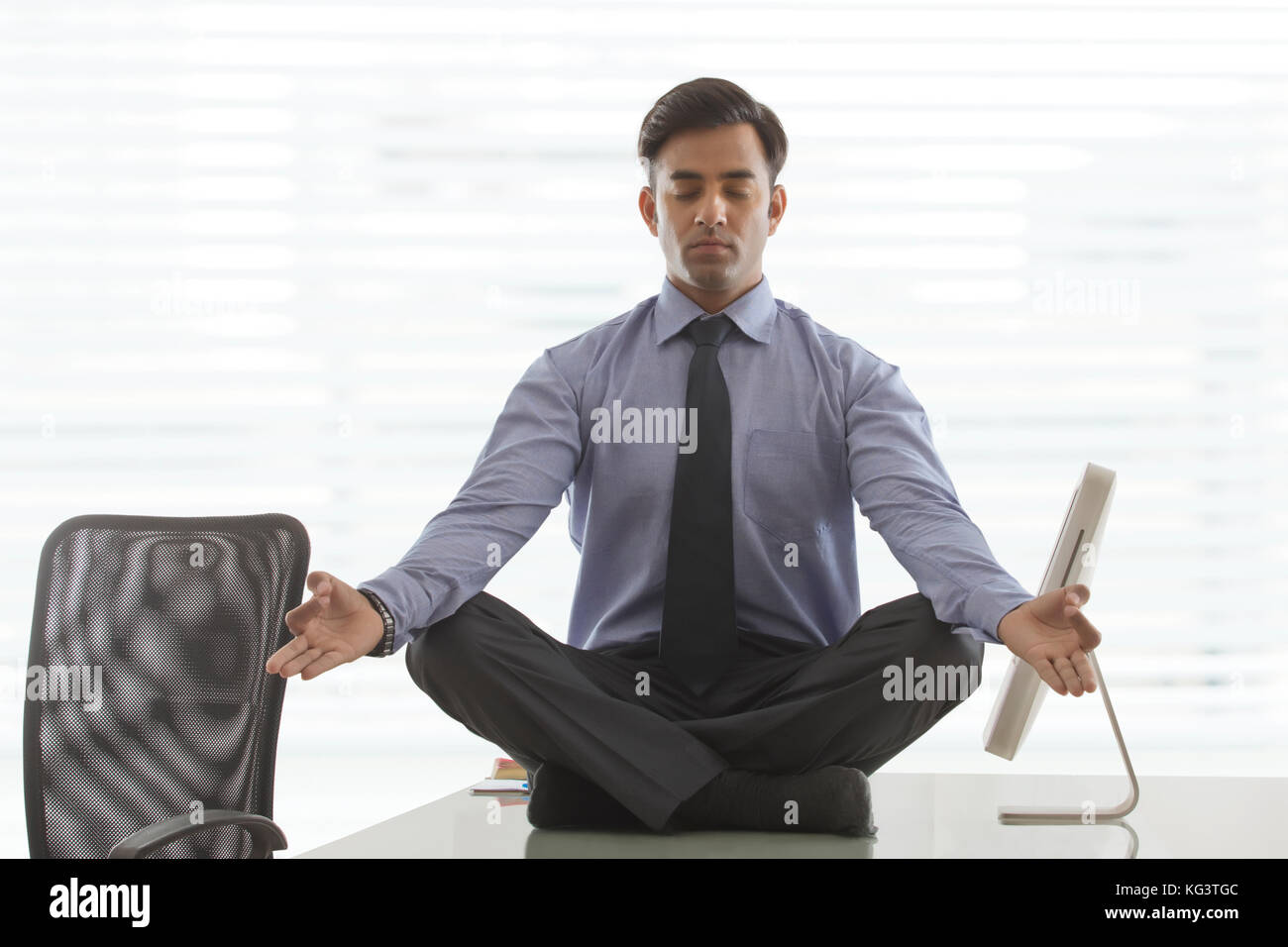 Young businessman sitting cross-legged doing yoga on desk in office Stock Photo