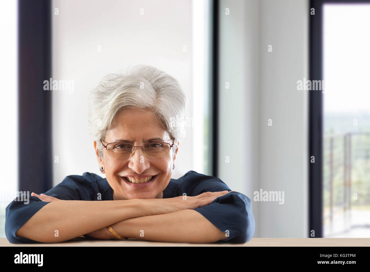 Smiling senior woman leaning on table arms crossed Stock Photo