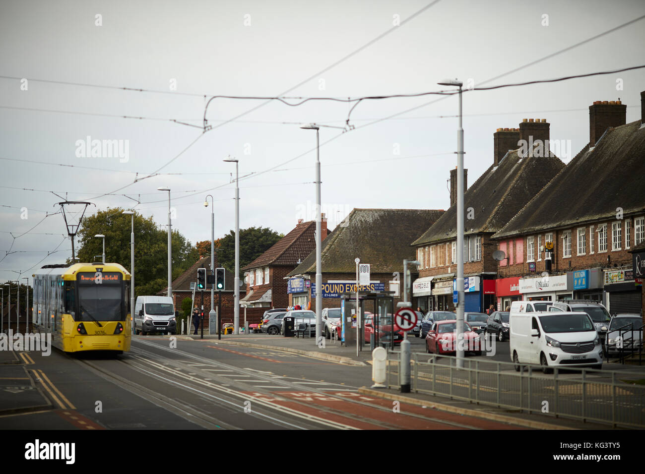 A yellow Metrolink Manchester tram at Hollyhedge Road, Benchill, Wythenshawe. passing a parade of independent local shops Stock Photo