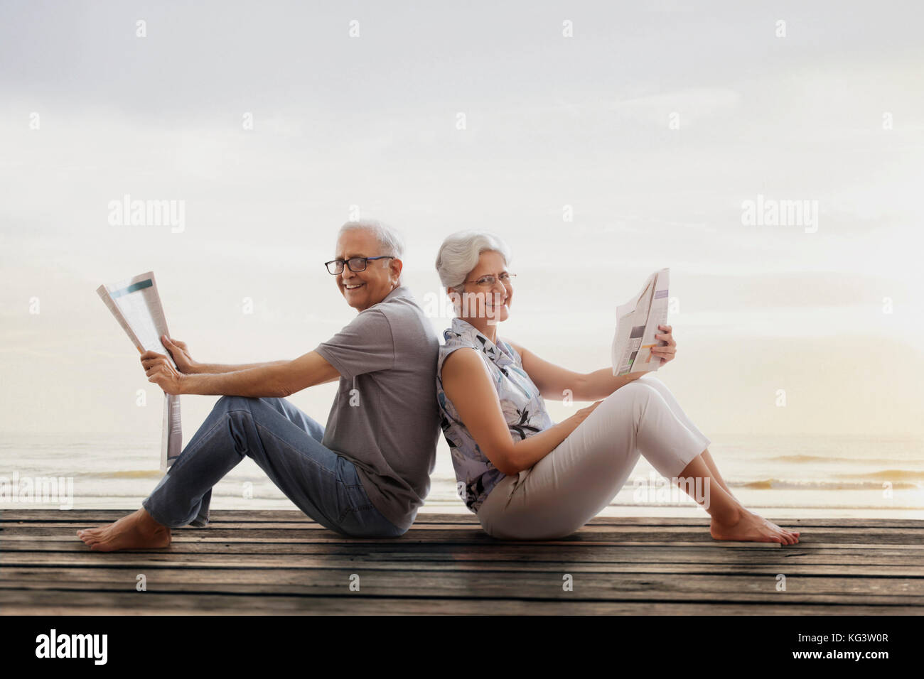 Senior couple sitting back to back on wooden dock near lake and reading newspaper Stock Photo