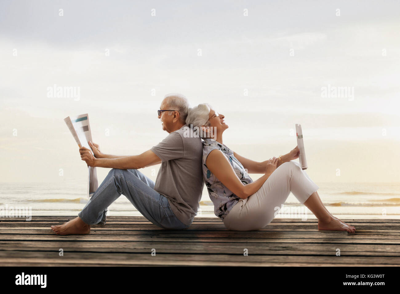 Senior couple sitting back to back on wooden dock near lake and reading newspaper Stock Photo