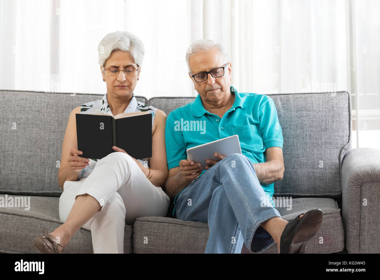 Senior woman reading book and senior man using digital tablet sitting on sofa Stock Photo