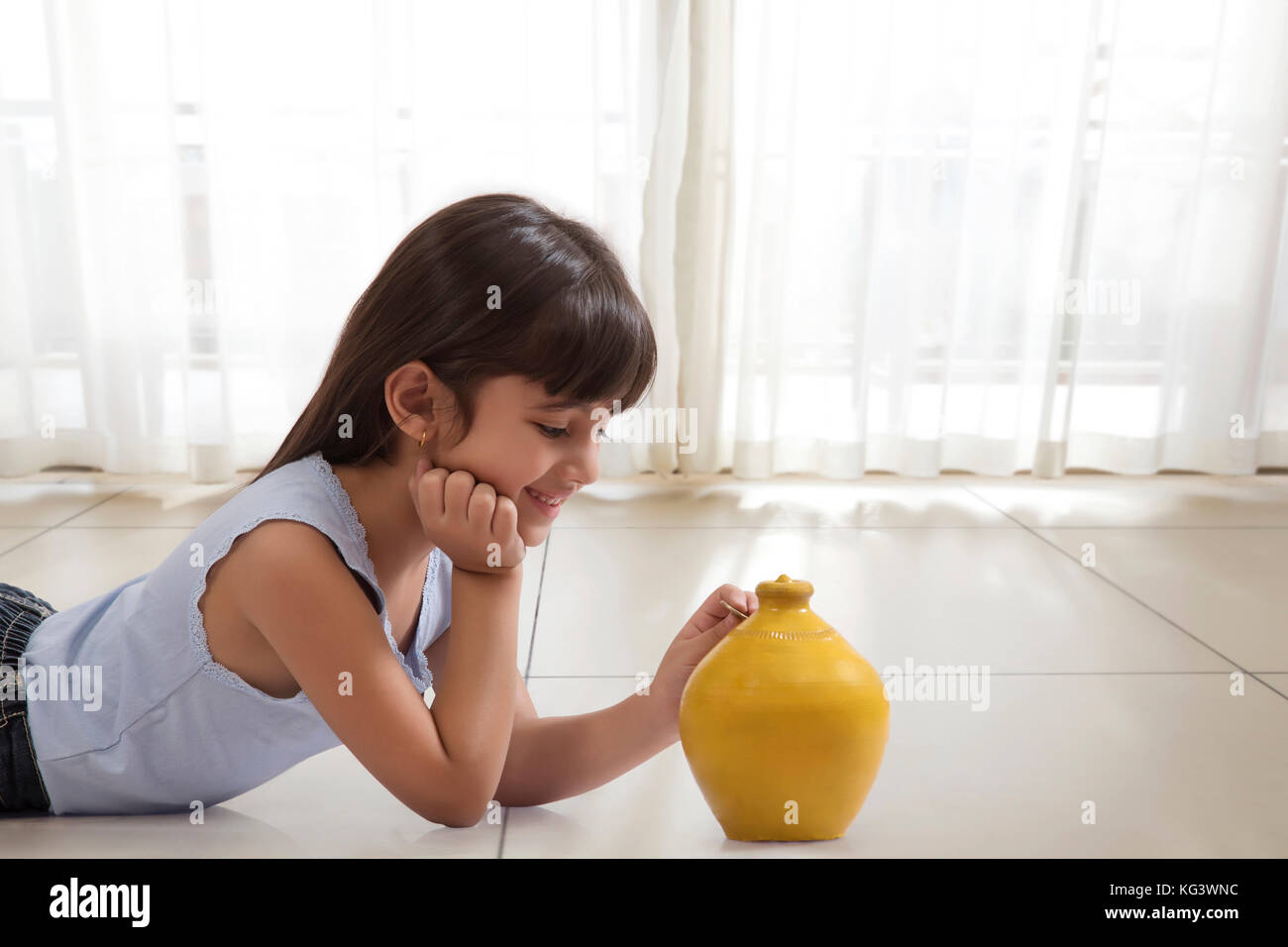 Little girl lying on floor and putting coin in clay piggy bank Stock Photo