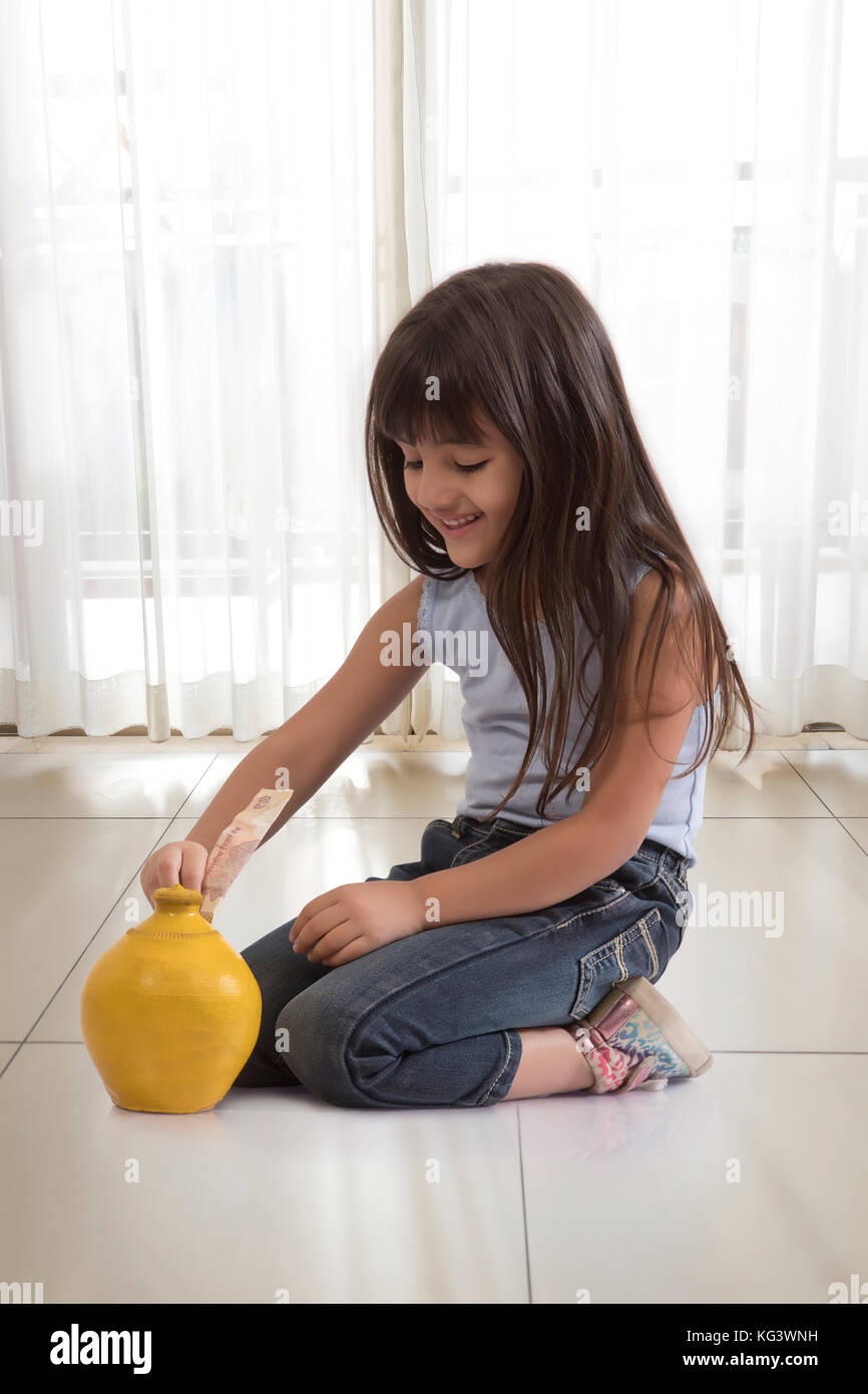 Little girl sitting on floor and putting bank note in clay piggy bank Stock Photo