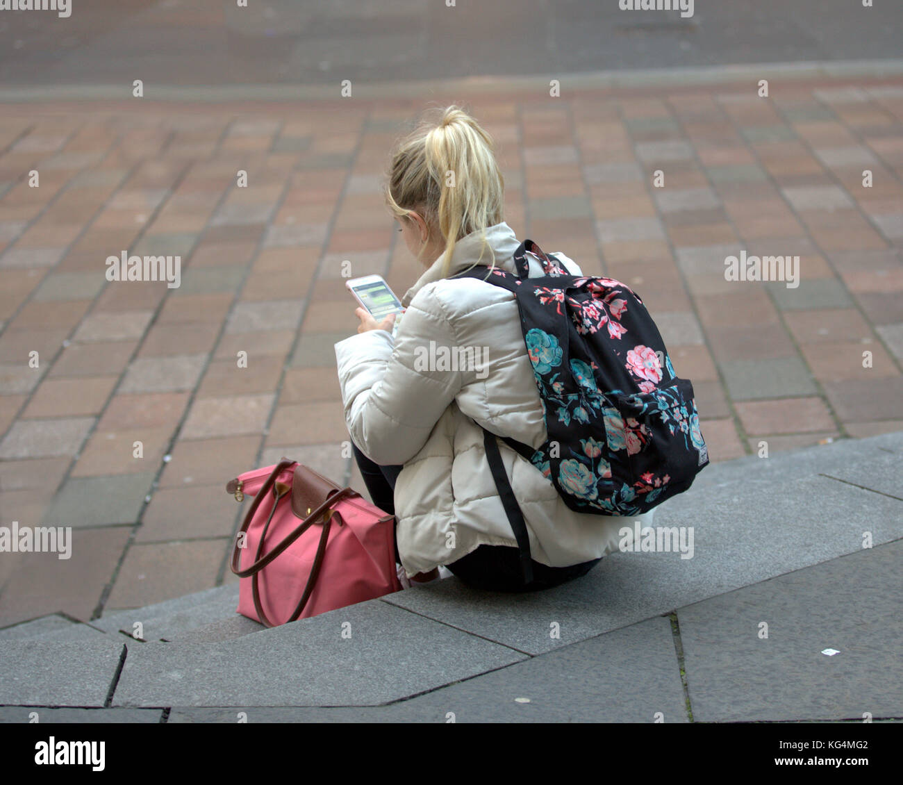 young colourful colorful girl student sitting on steps with smartphone and a backpack viewed from behind Stock Photo