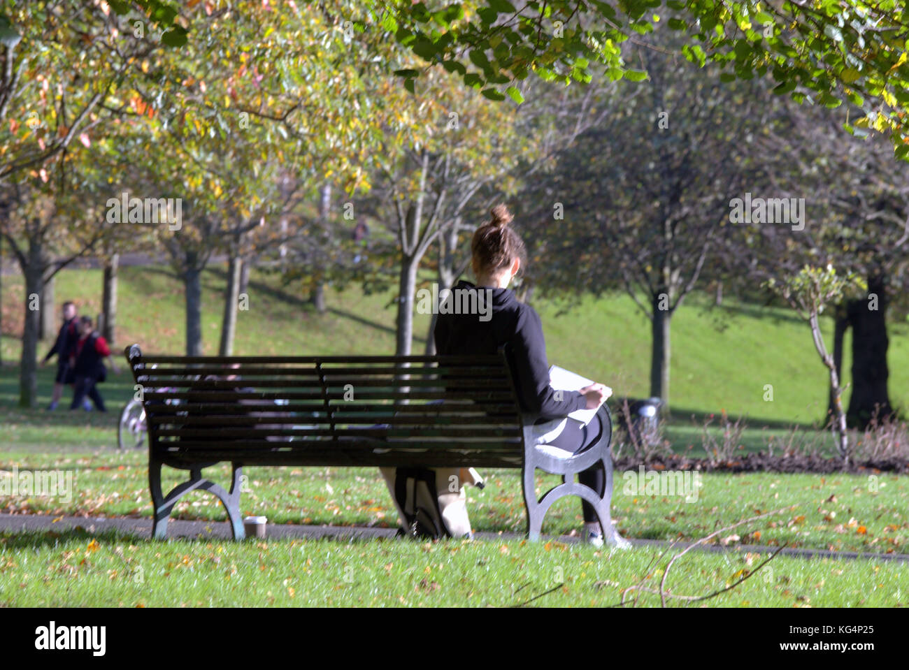 young girl student studying in Kelvingrove Park  sunny day sitting on bench with people in the background viewed from behind Stock Photo