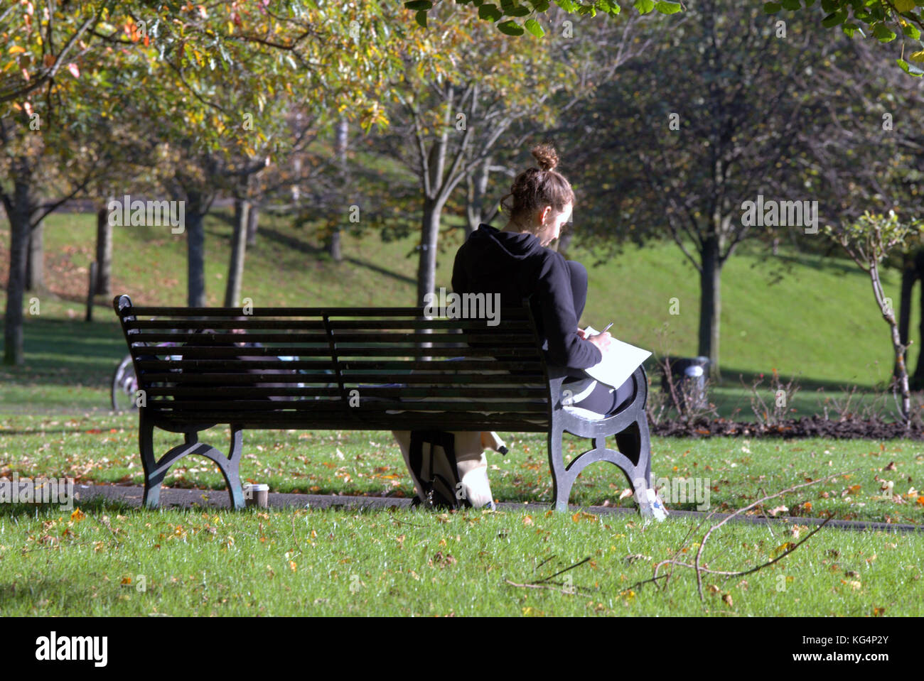 young girl student studying in Kelvingrove Park sunny day sitting on bench with  viewed from behind Stock Photo