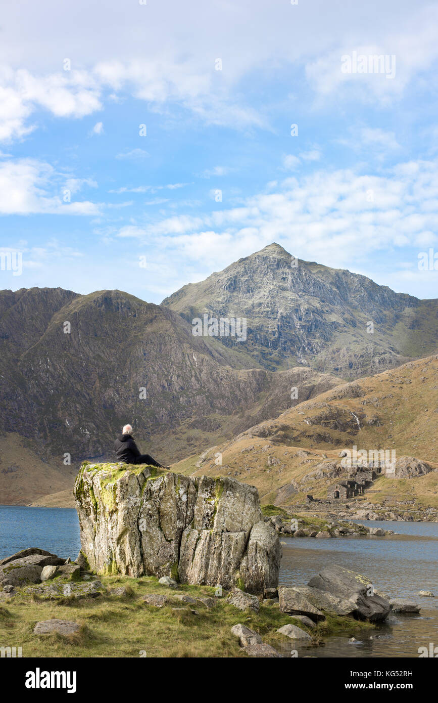 Solitary white-haired man sits on a rock gazes at the peak of mount Snowdon, the highest mountain in England and Wales. Stock Photo