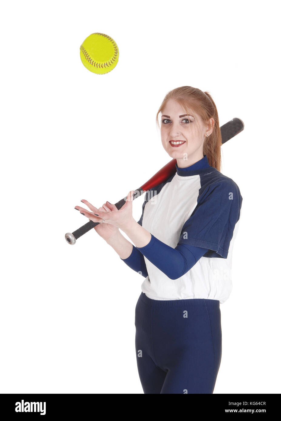 A smiling young woman standing in her blue softball uniform playing with the yellow ball holding her bat, isolated for white background Stock Photo
