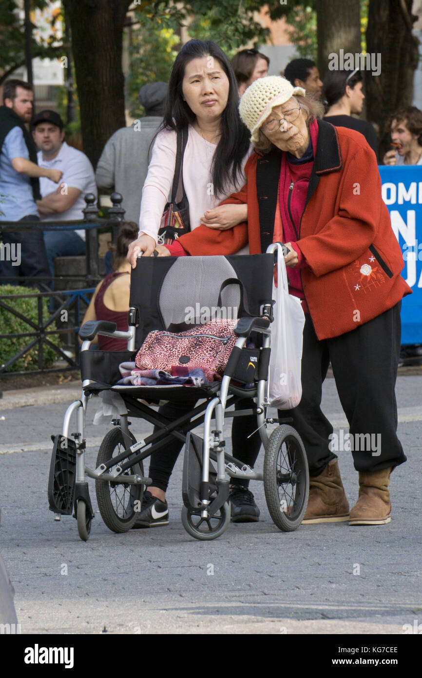 A young Asian American woman accompanies what might be her grandmother for a walk outside of her wheenchair. In Union Square Park in New York City. Stock Photo