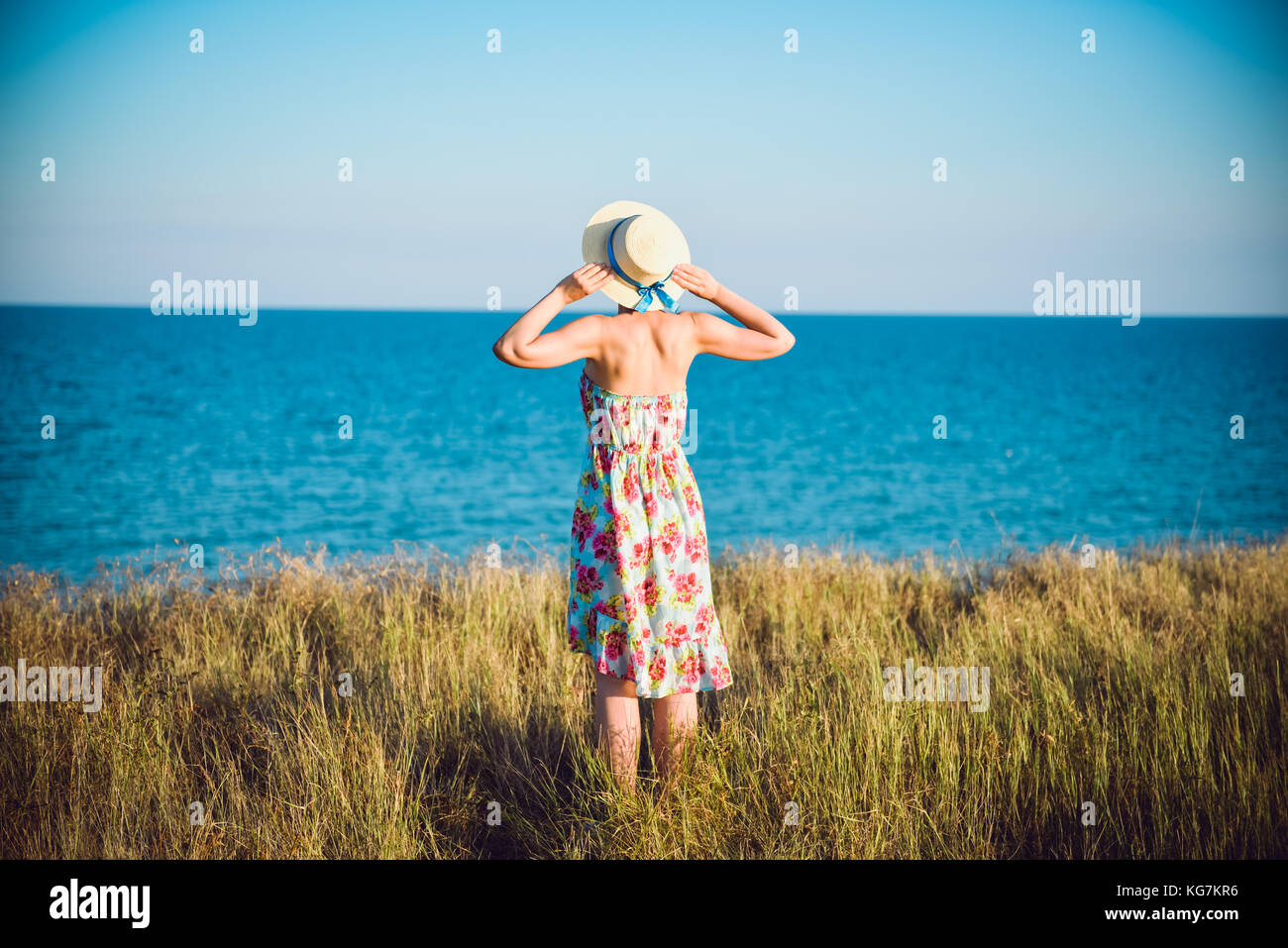 Summer portrait of young pretty woman in a straw hat and dress standing backwards in the grass and looking to the sea. Girl enjoy nature sea paradise  Stock Photo