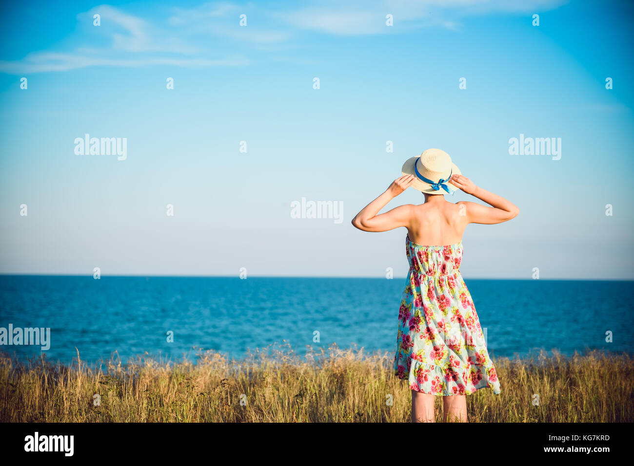Summer portrait of young pretty woman in a straw hat and dress standing backwards in the grass and looking to the sea. Girl enjoy nature sea paradise  Stock Photo