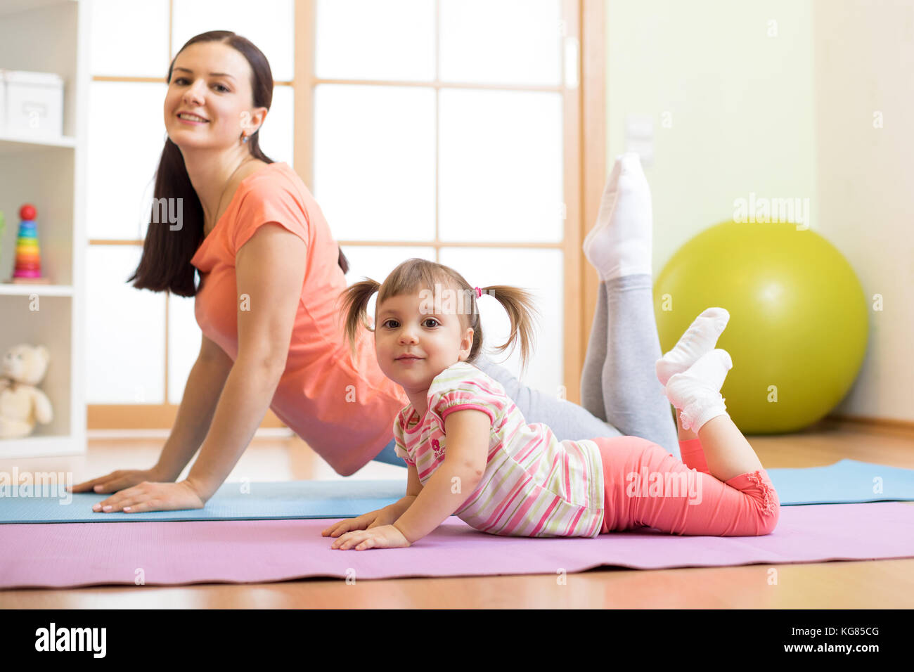Mother and child daughter doing yoga exercises on floor in room at home. Family having fun indoors with fitness. Stock Photo