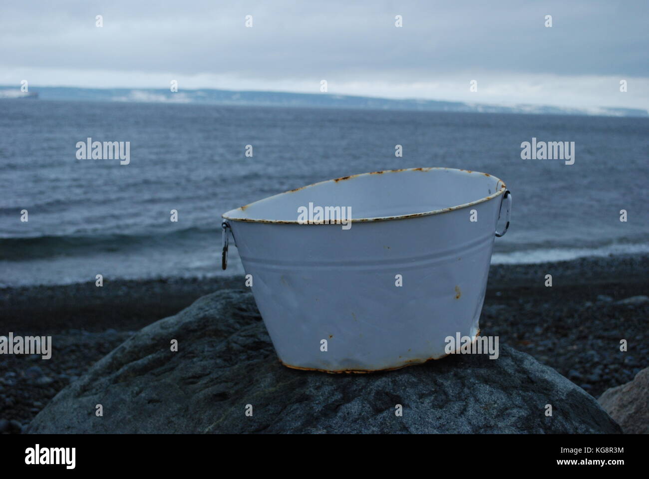 White metal bucket sitting atop a rock, on a beach, winter. Conception Bay South, Newfoundland and Labrador, Canada Stock Photo
