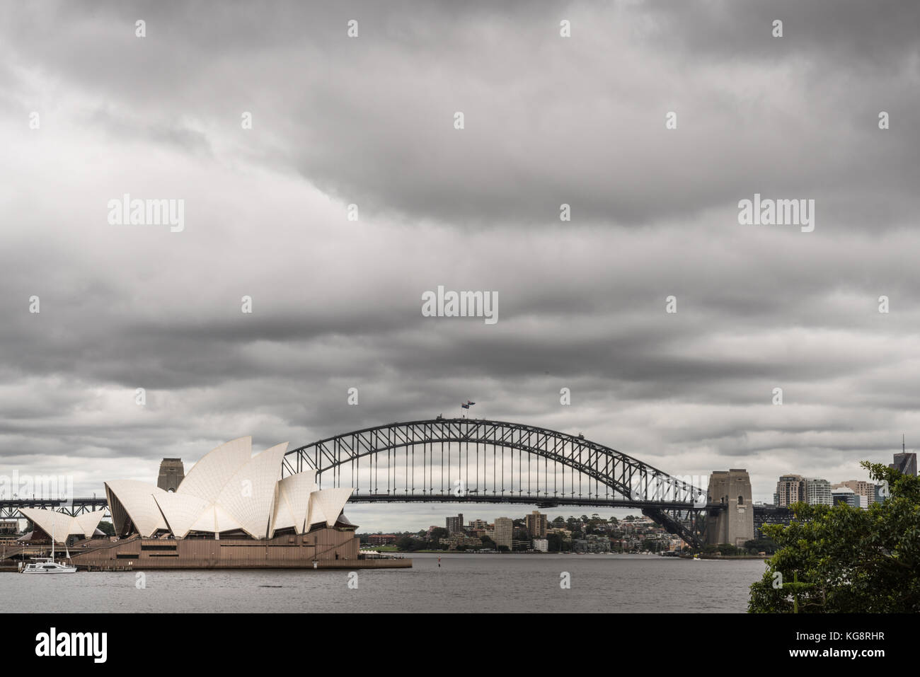 Sydney, Australia - March 23, 2017: White Opera House and black bow of Harbour Bridge behind under heavy rainy cloudscape. Kirribilli tall buildings i Stock Photo