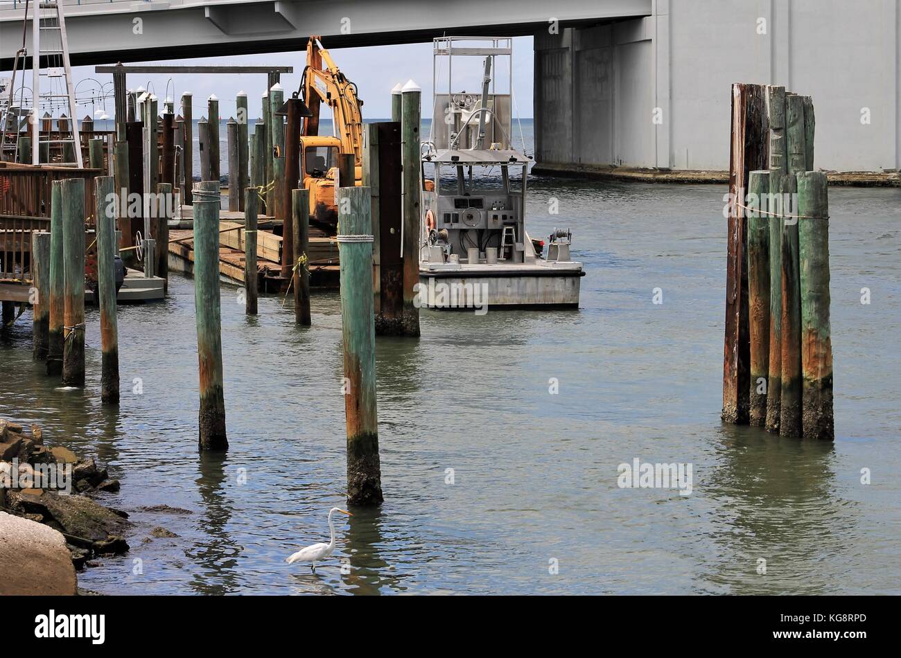 Boat, and barge with excavator dock side at a pier in Treasure Island, Florida. Pilings visible sticking out of the water. Lift bridge in background Stock Photo