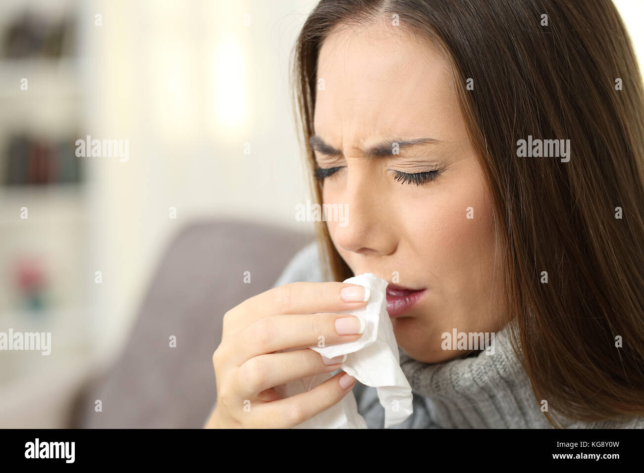 Ill woman coughing using a tissue sitting on a couch in the living room