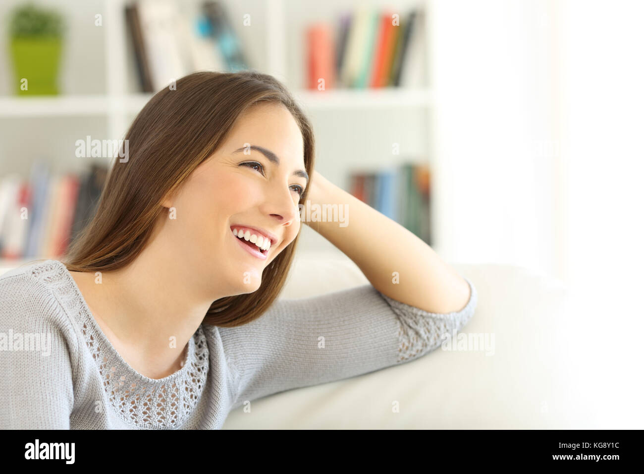 Portrait of a happy pensive girl looking away sitting on a sofa in the living room at home with copy space in white Stock Photo