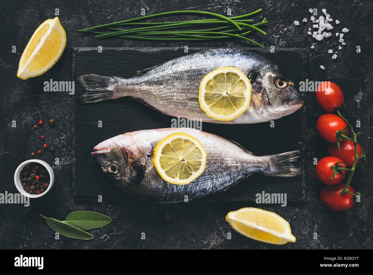 Fresh sea beam or dorado fish, lemon, herbs, spices and vegetables on black stone slate background. Top view. Fresh fish ready for cooking. Toned imag Stock Photo