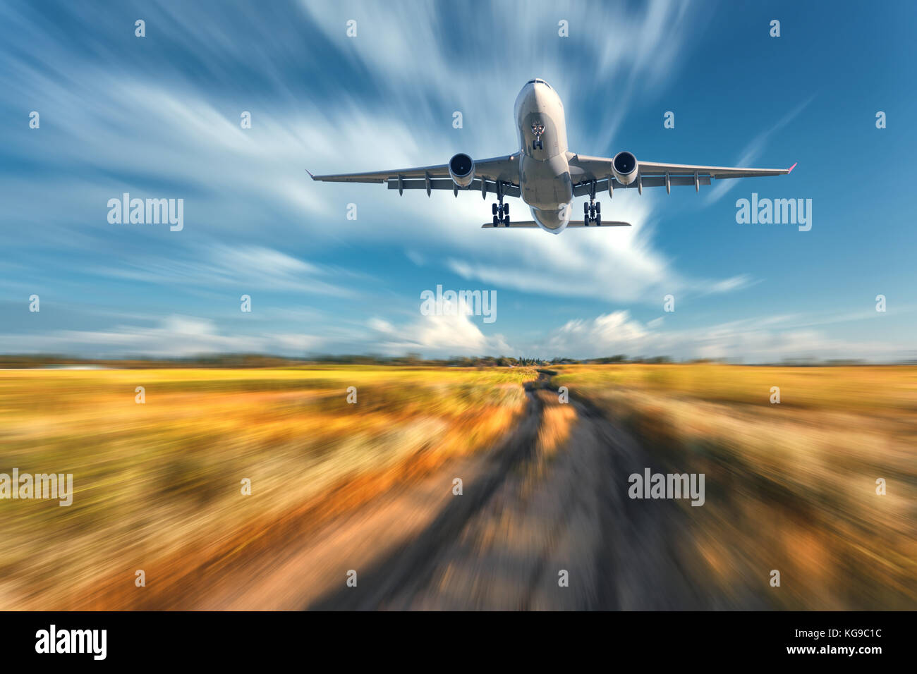 Airplane with motion blur effect. Landscape with flying passenger airplane and blurred blue sky with clouds, orange grass field with trail at sunset.  Stock Photo