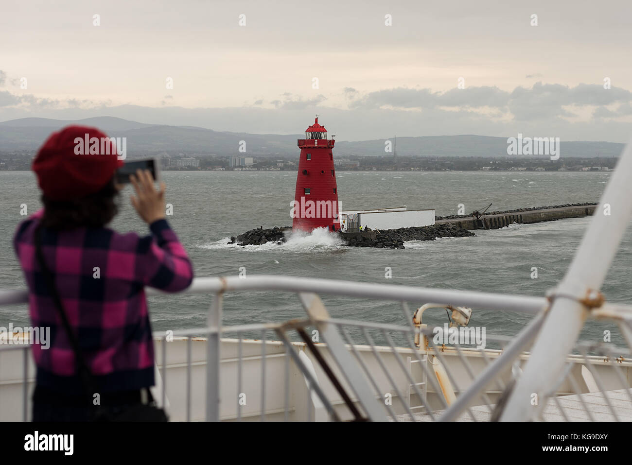 A women with a red hat takes a smart phone snap shop holiday photo of Poolbeg lighthouse out on deck of a ferry leaving Dublin port. Stock Photo