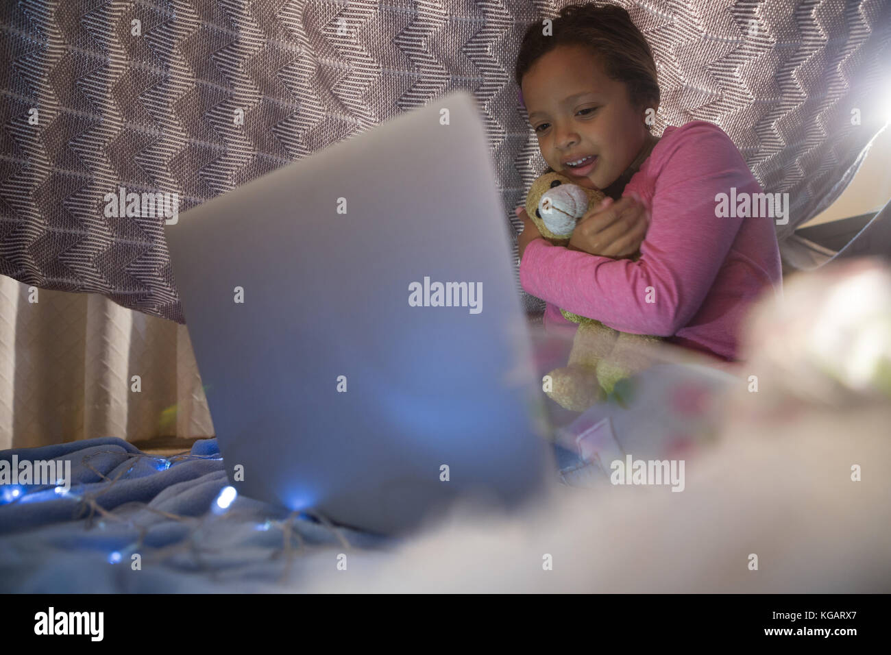 Girl holding teddy bear and looking at laptop in bedroom Stock Photo