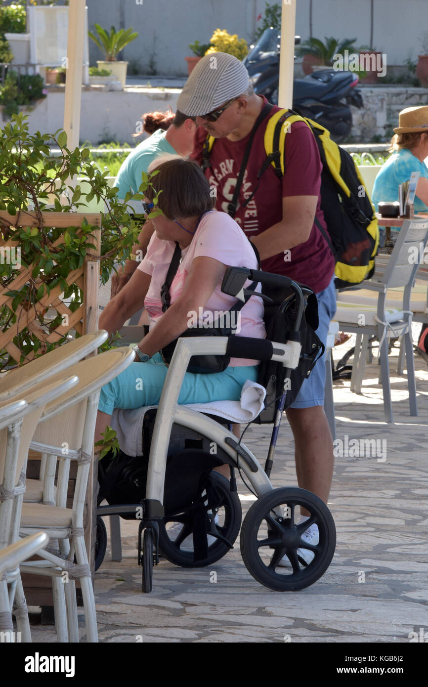 a disabled woman or lady on holiday in Greece being pushed in a wheelchair by her partner, carer or husband. disable access in wheelchairs on holiday. Stock Photo