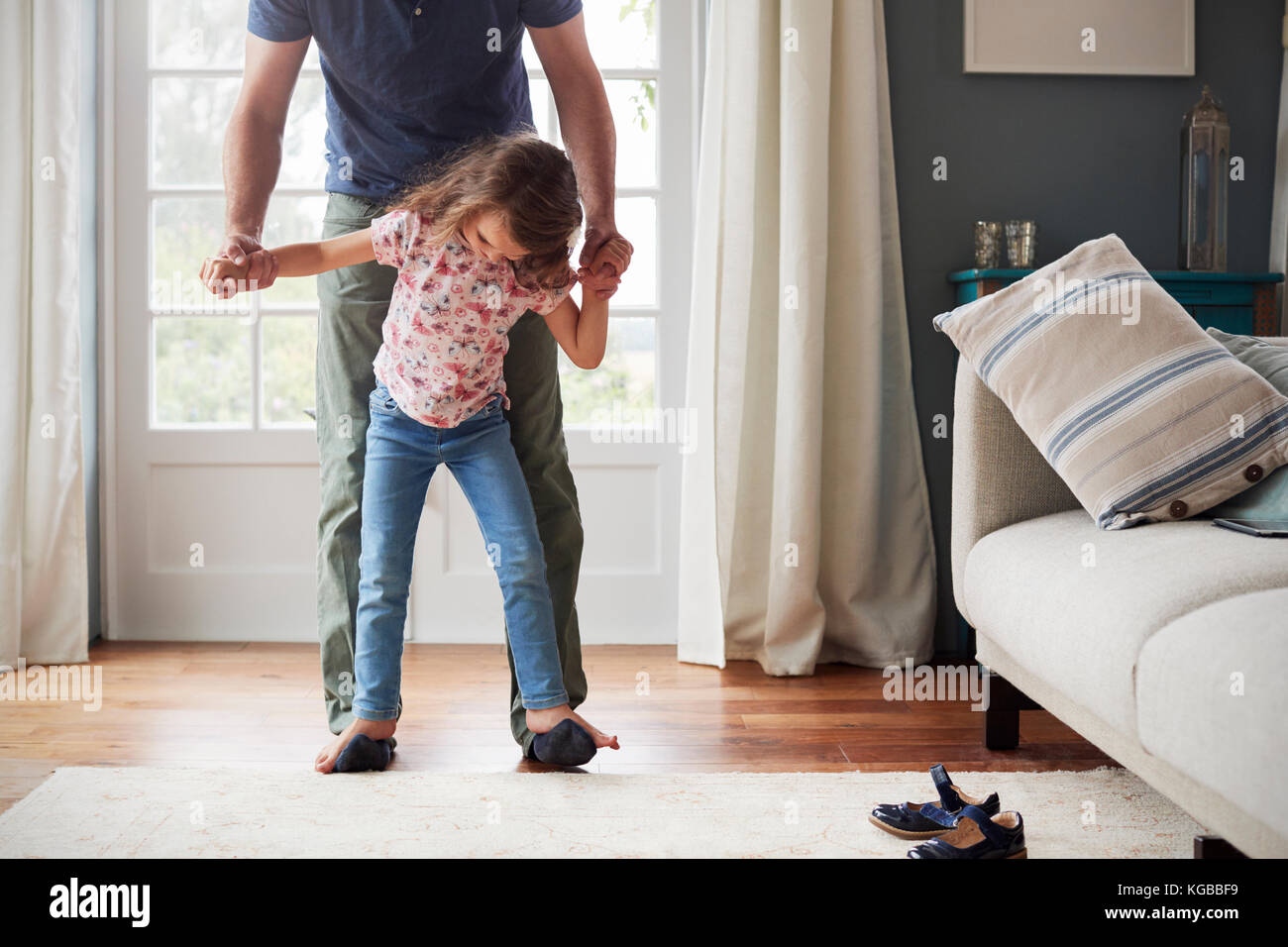 Girl balances walking on father’s feet at home, looking down Stock Photo