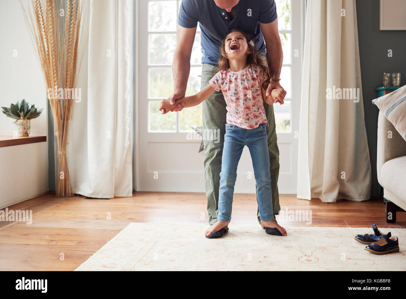 Girl balancing on father’s feet at home, looking up Stock Photo