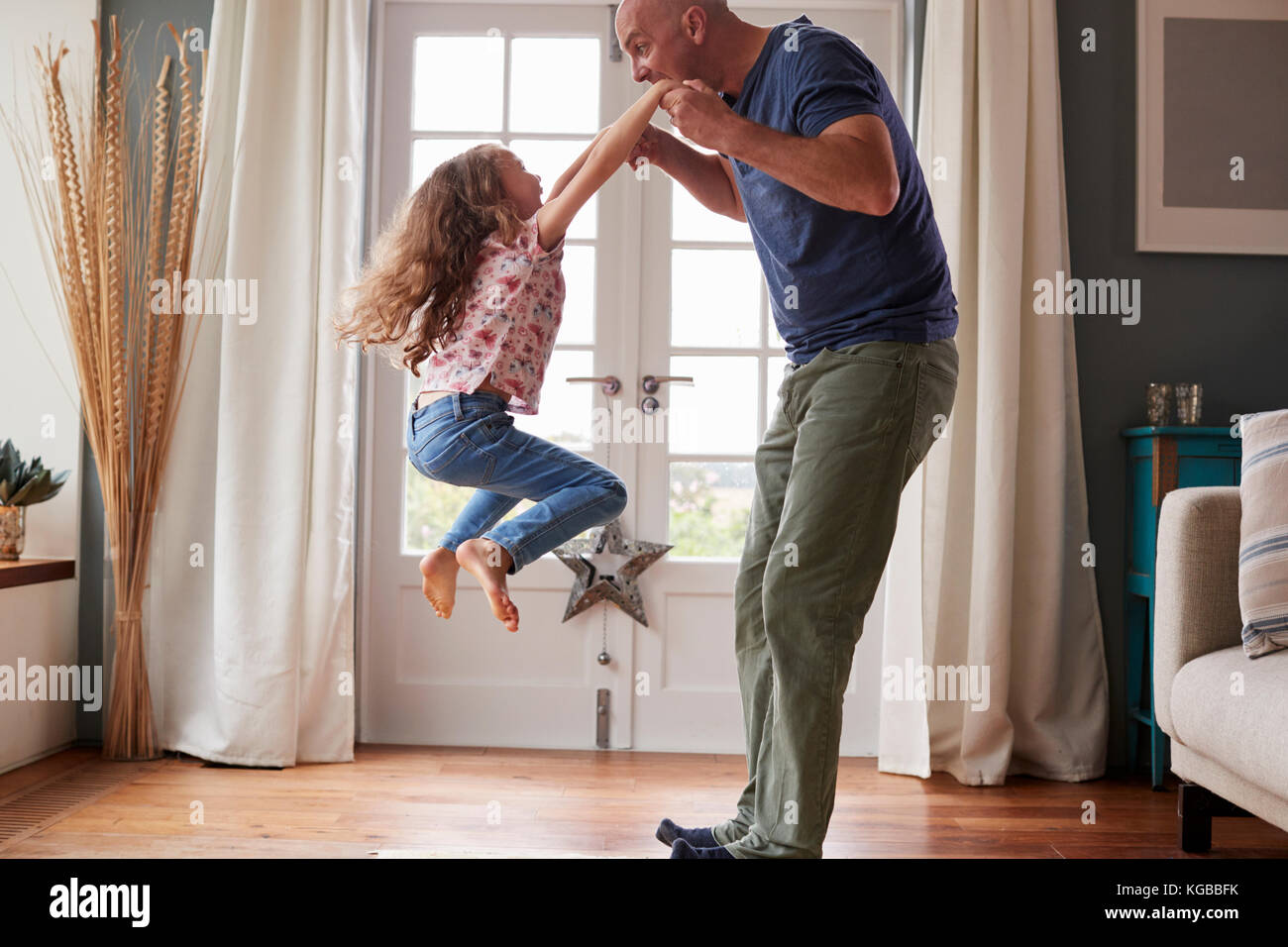 Girl jumping in the air at home holding dad’s hands Stock Photo