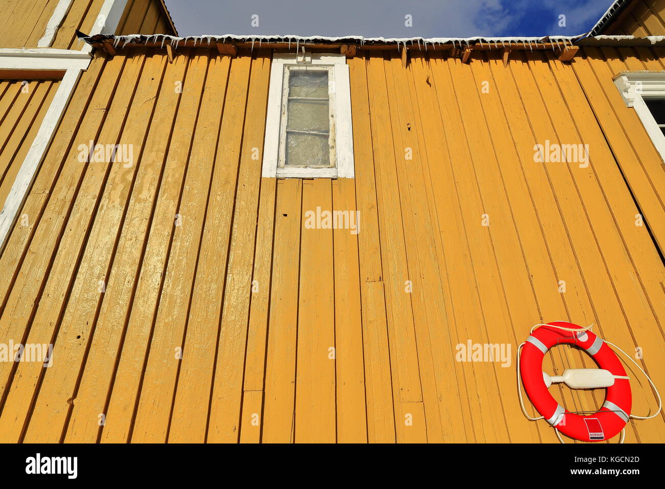 Yellow facade of fishing port warehouse-closed old window-icicles hanging from roof gutters-red round life saver hanging on the wall-white windowframe Stock Photo