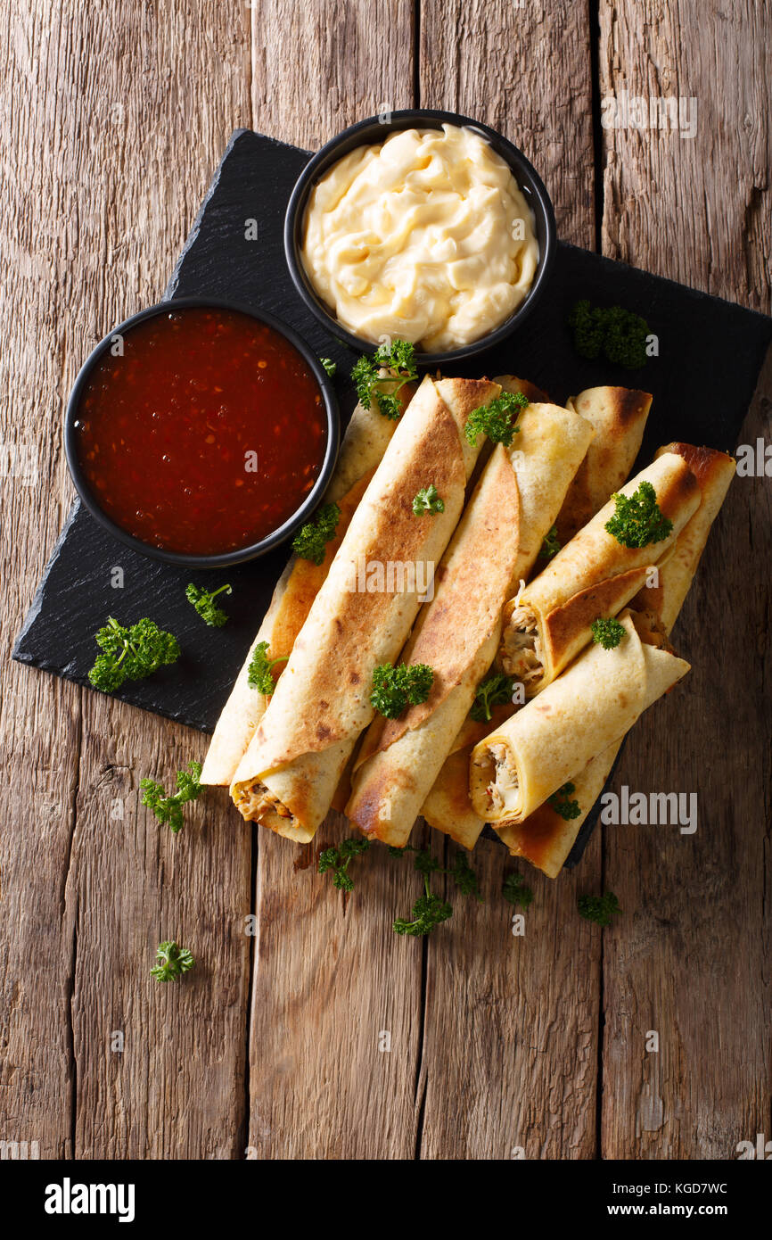 Mexican taquitos with chicken and cheese stuffing close-up, and sauces on the table. Vertical top view from above Stock Photo