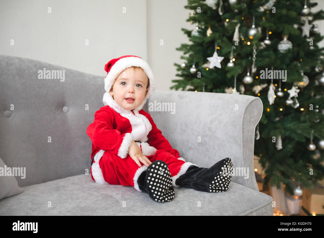 Baby in santa suit sit on sofa with Christmas tree Stock Photo