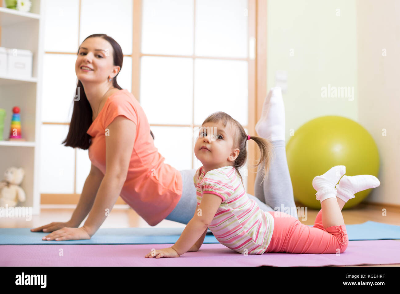Mother and kid daughter doing yoga exercises on floor in room at home. Family having fun indoors with fitness. Stock Photo