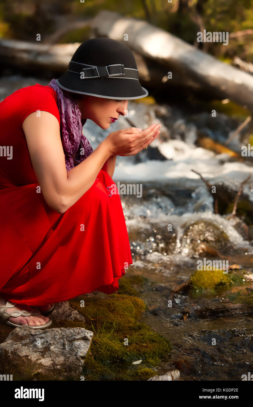A young woman crouches to drink from a mountain stream Stock Photo