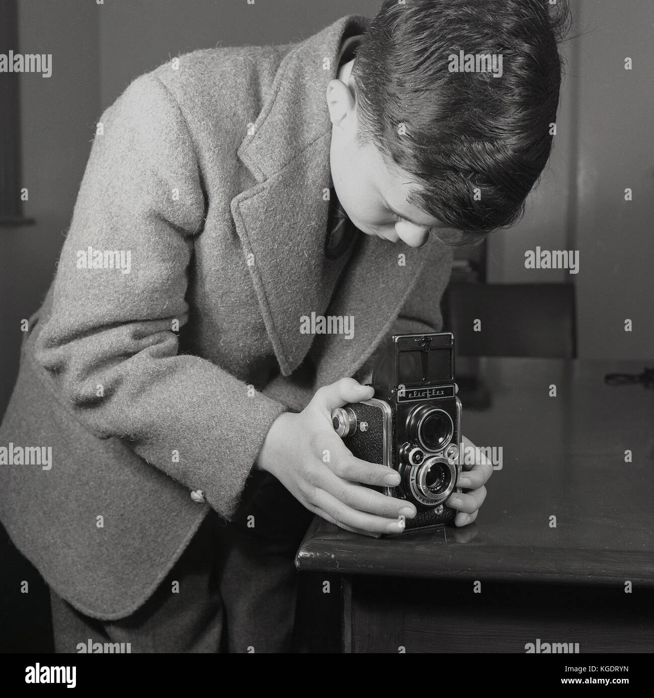 1950s, historical, England, Uk, a smartly dressed teenage boy wearing a jacket and tie uses a table corner for stability as he takes a picture using a Ferrania Elioflex camera. This was an Italian made film camera that imitated the look of a TLR ( Twins Lens Reflex) Rolleiflex, which were the professional cameras of their day, but this make was not one as the viewfinder was a not a true reflex finder. It was known therefore as a 'pseudo TLR' but was popular in this era. Stock Photo