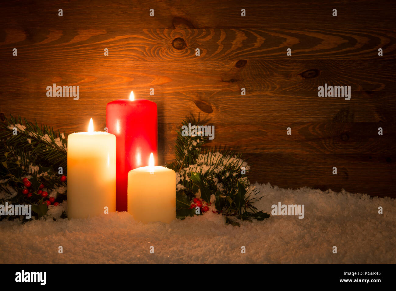 Christmas candles with Holly and conifer branches on an artificial snow and wooden background. Stock Photo