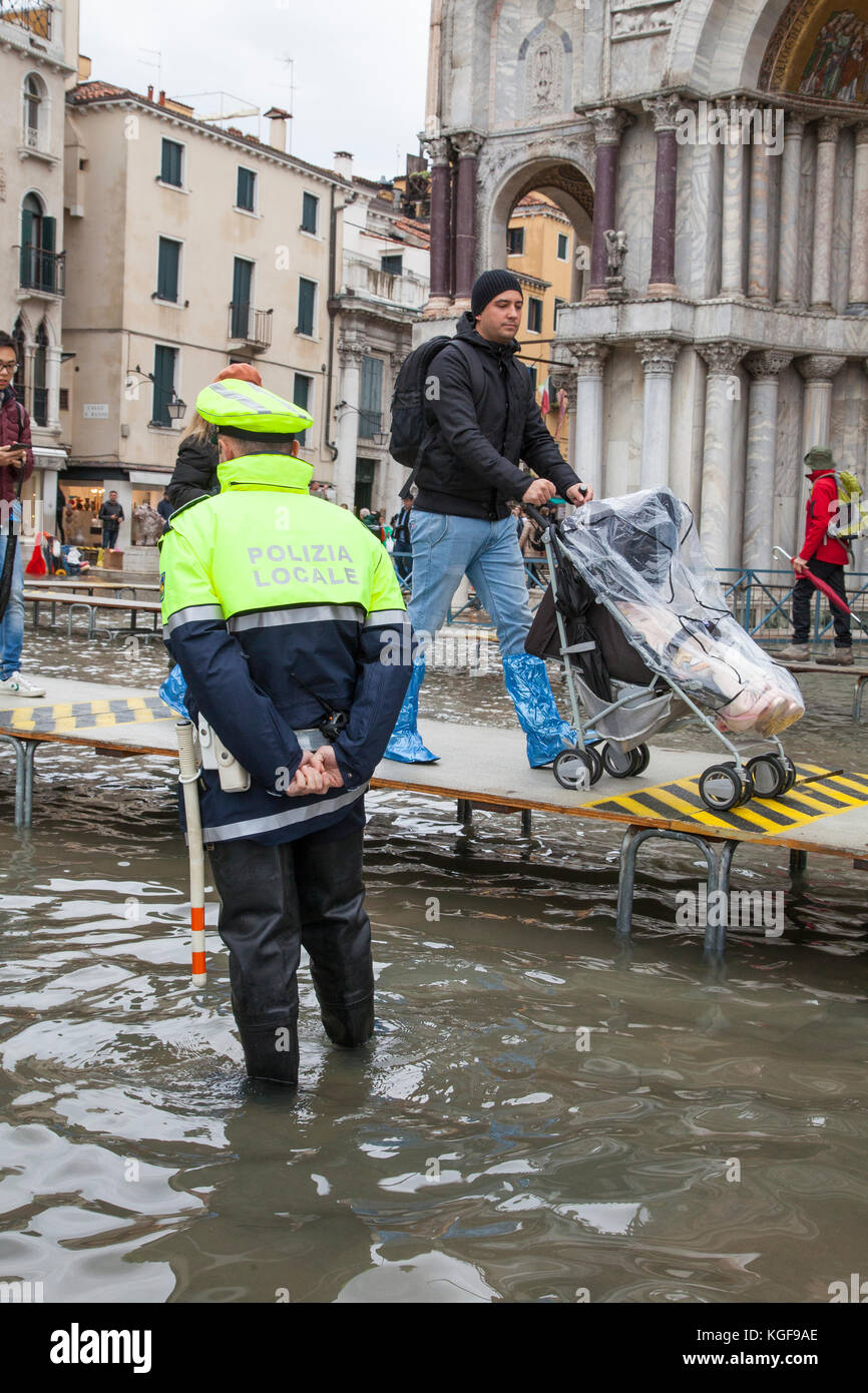 Venice, Veneto, Italy. 7th Nov, 2017. Acqua Alta high tide of 115cm from the lagoon causing temporary flooding in Piazza San Marco. Passarelle, or elevated walkways, are installed for pedestrian traffic with a father pushing his young child in a push chair covered in plastic past a Polizia Locale officer. He is wearing disposable plastic shoe covers. Credit: Mary Clarke/Alamy Live News Stock Photo