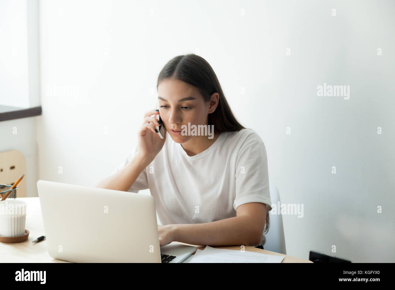 Attentive young adult woman check her order in laptop computer and confirms details by smartphone. Corporate employee planning her vacation. Stock Photo