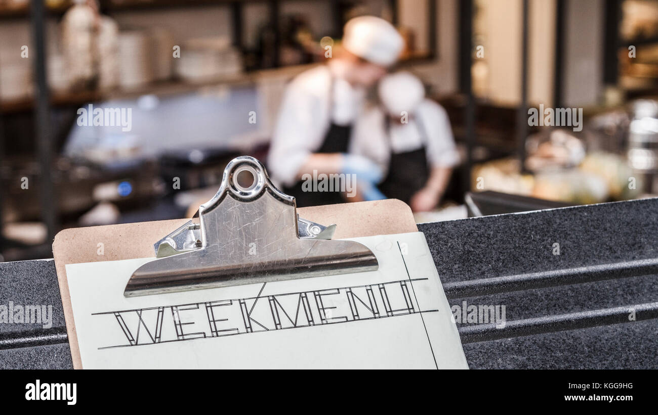 Week menu clipboard with restaurant kitchen and two cooks busy in the background. Stock Photo