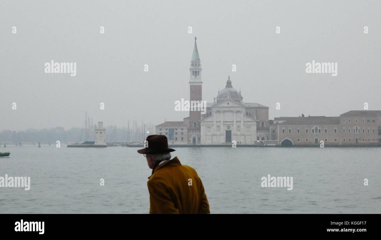 Dapper elderly man in mustard yellow coat and trilby hat walks alone, opposite the church of Saint Giorgio Maggiore off the shores of Venice, Italy Stock Photo
