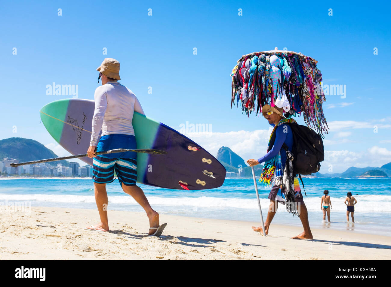 RIO DE JANEIRO - MARCH 21, 2017: Brazilian vendor selling bikinis walks alongside a man carrying his stand-up paddle surfboard along Copacabana Beach. Stock Photo