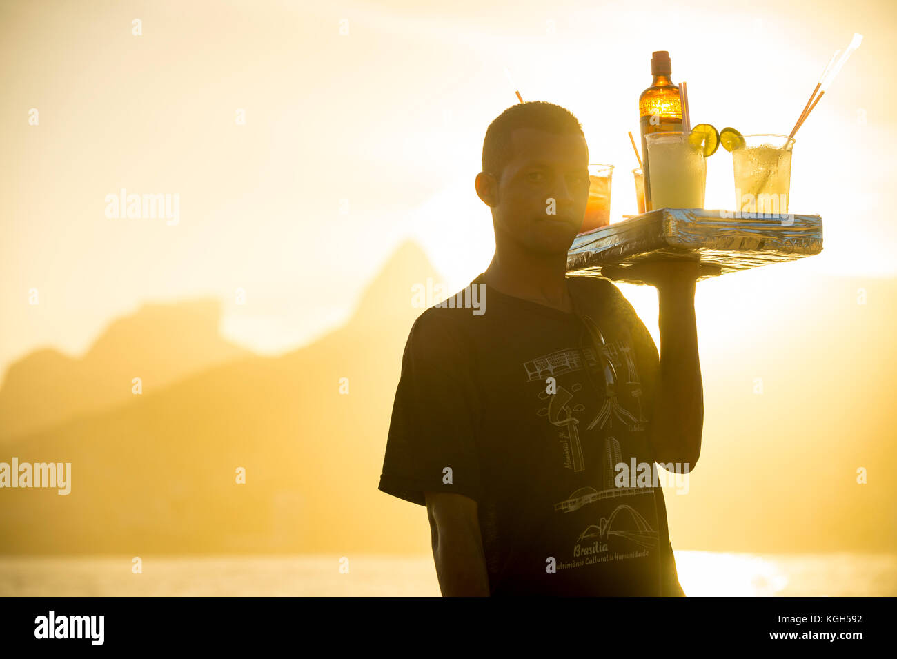 RIO DE JANEIRO - MARCH 20, 2017: Sunset silhouette of young man selling caipirinha cocktails at Arpoador with Two Brothers mountain in the background. Stock Photo
